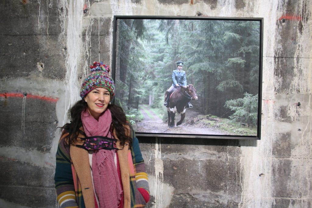 Die Künstlerin Sandra Mann vor dem Farbfoto „Bulle auf Kuh“ Model Julia reitet auf der Kuh „Perle“ durch den Bayrischen Wald. Julia trägt dazu eine DDR-Polizeiuniform Fotograf: Edda Rössler