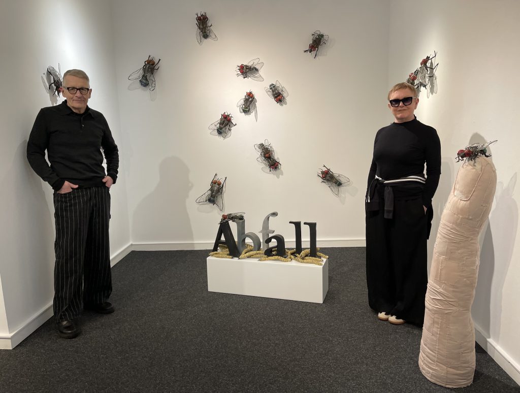 Ingrid und Andreas Honneth vor Fliegen, Abfall, Maden Ekel kippt ins Ornament: Überdimensionierte Fliegen umschwirren das Wort „Abfall“, das wie eine Reliquie präsentiert wird. Vanitas als Montage aus Konsum, Verfall und Ironie. Foto: Edda Rössler