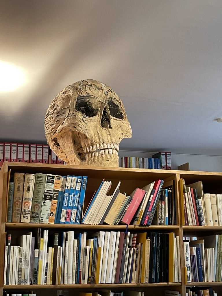 Ingrid Honneth: Totenschädel auf Bücherregal Memento im Lesesaal: Ein aus Papier modellierter Schädel blickt über Bücher hinweg. Vergänglichkeit sitzt hier buchstäblich auf dem Wissen. Foto: Edda Rössler